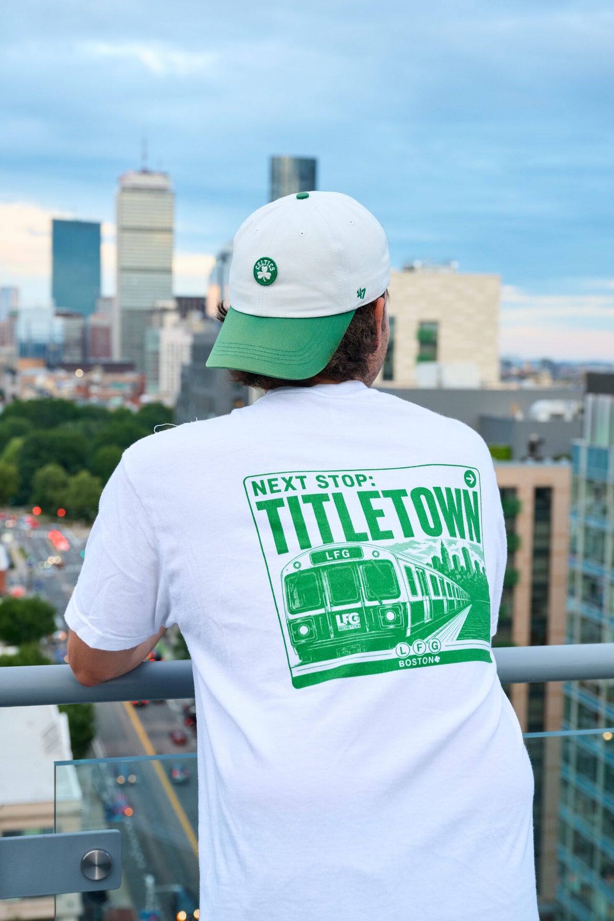 fan in the lfg greenline pocket tee, next stop titletown back print, with the boston skyline behind